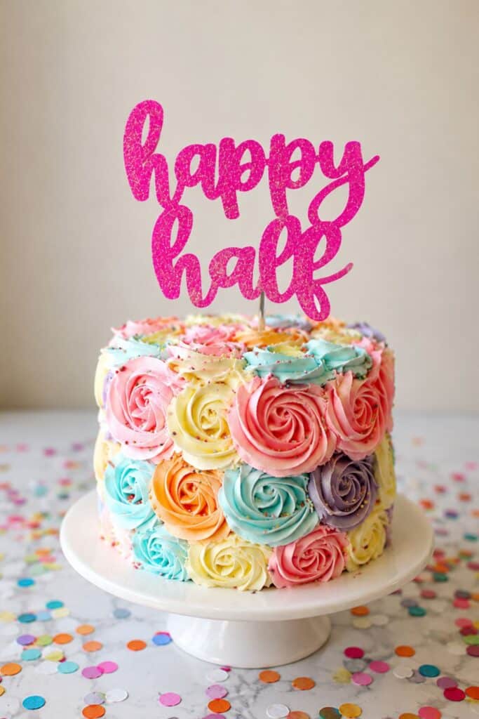 Colorful rosette half birthday cake with bright pastel frosting swirls and glitter “happy half” topper for a fun 6 month birthday party idea.