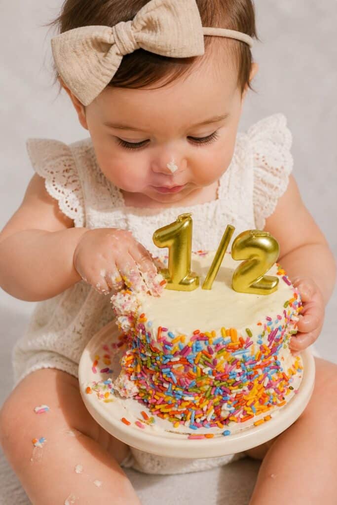Baby girl enjoying a colorful sprinkle half birthday smash cake with gold 1/2 candles for a sweet and simple 6 month birthday photoshoot idea.
