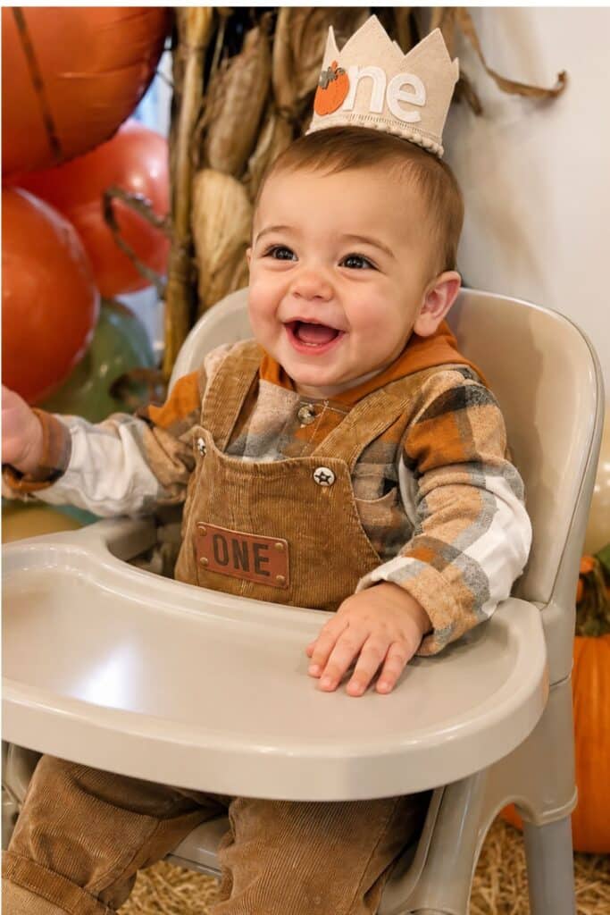 Pumpkin patch first birthday party backdrop with fall balloon garland, hay bale, pumpkins, mums, and rustic wood sign.