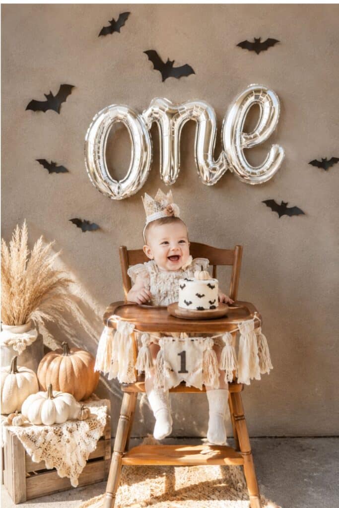 Neutral Halloween first birthday cake smash setup with silver one balloon, black bats, white pumpkins, and baby girl in a high chair.
