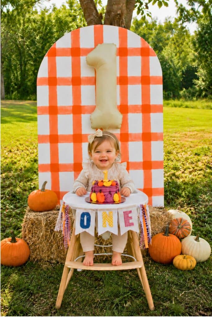 Pumpkin first birthday cake smash setup with orange gingham backdrop, hay bales, pumpkins, one balloon, and baby girl in a high chair.
