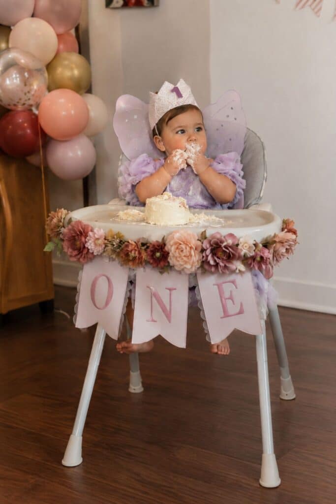 Fairy first birthday cake smash with baby girl in purple fairy wings, floral high chair banner, pink balloons, and one birthday crown.